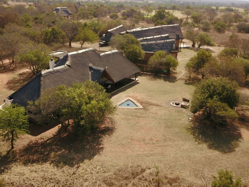 Aerial view of a lodge property featuring multiple thatched-roof buildings, a swimming pool, and a fire pit set within a natural bushveld landscape.