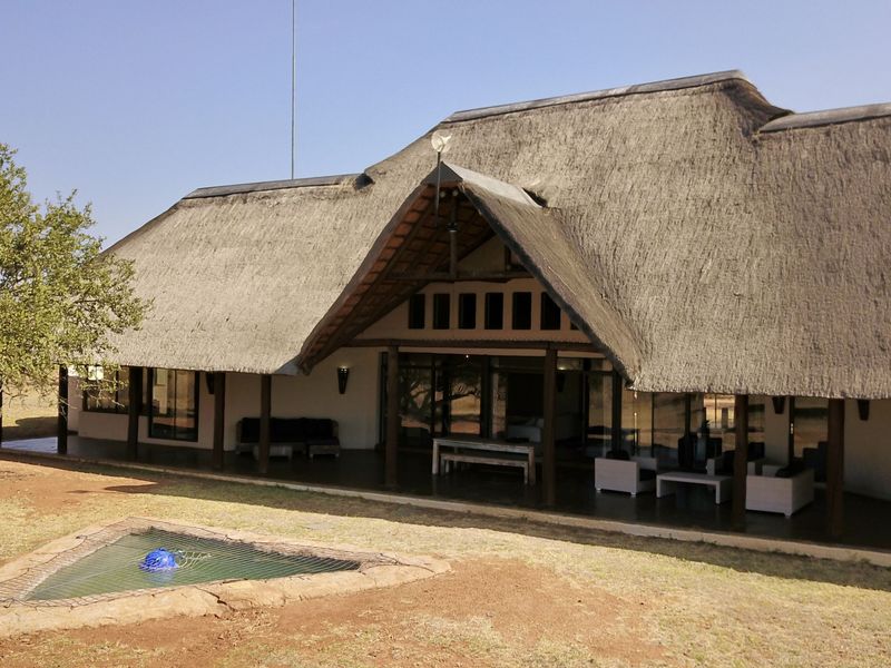 Rear view of a lodge featuring a traditional thatched roof, expansive covered patio, and large sliding glass doors overlooking the natural landscape.