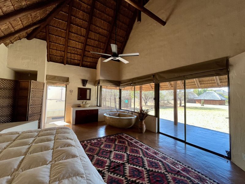 Spacious lodge bedroom featuring a high thatched ceiling, exposed timber beams, an integrated stone bathtub, and large sliding doors opening to the outdoor area.