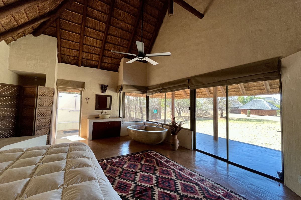 Spacious lodge bedroom featuring a high thatched ceiling, exposed timber beams, an integrated stone bathtub, and large sliding doors opening to the outdoor area.