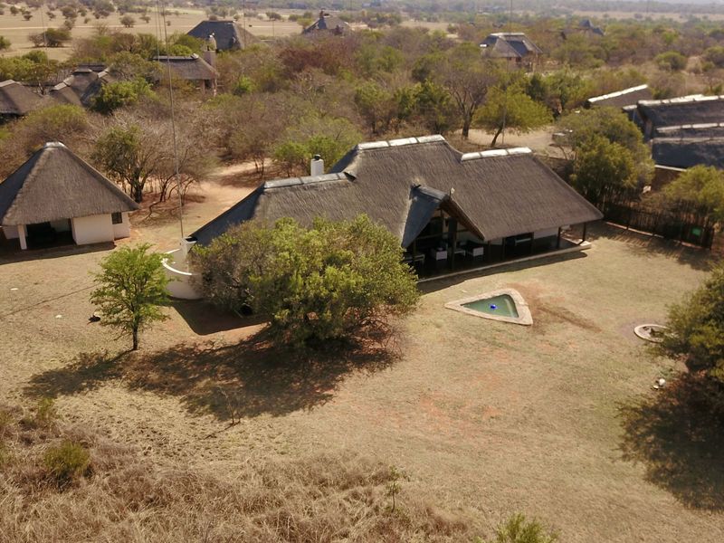 Aerial view of a lodge property featuring multiple thatched-roof buildings, a swimming pool, and surrounding natural bushveld landscape.