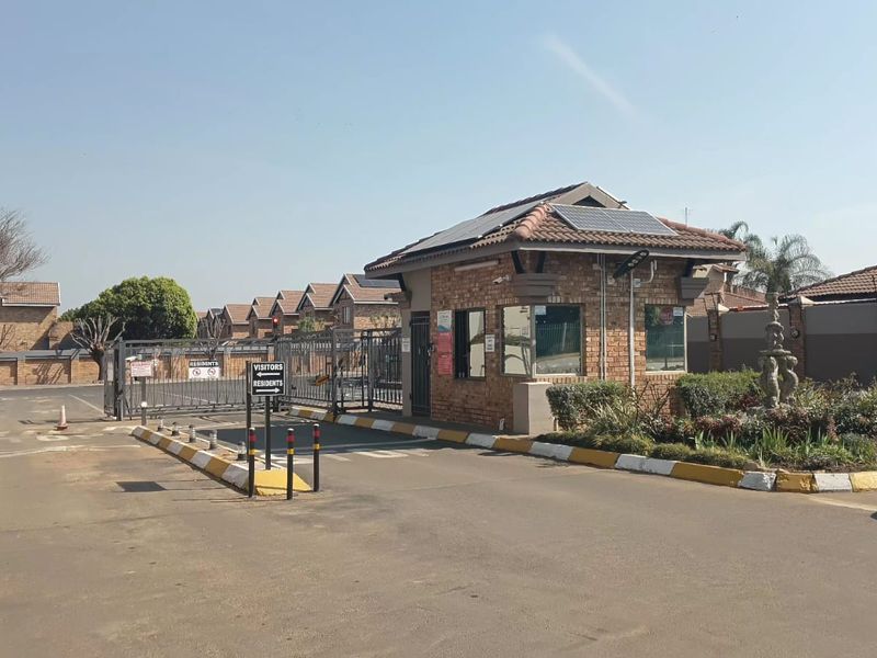 The entrance to the apartment complex features a secure gated access point with a brick guard house, solar panels, and well-maintained perimeter fencing.