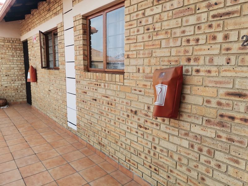 Exterior view of an apartment entrance featuring a face brick facade, tiled walkway, and timber-framed windows with security bars.