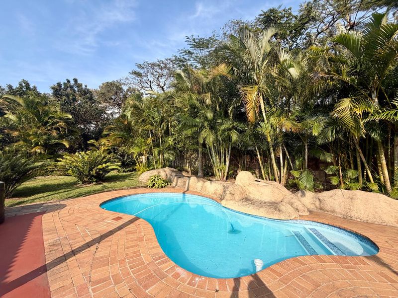A well-maintained swimming pool area featuring natural rock landscaping, brick paving, and lush surrounding greenery.