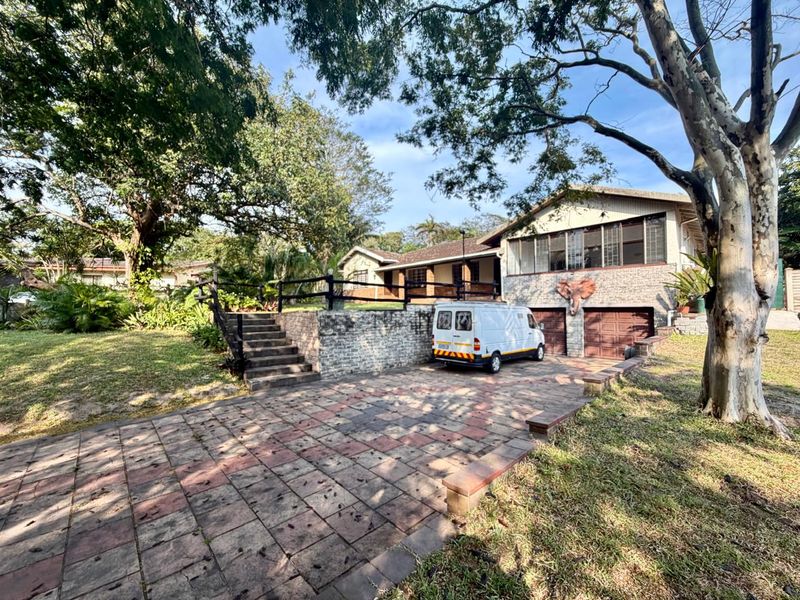 Front elevation of a residential property featuring a paved driveway, double garage, and elevated garden area with mature trees.