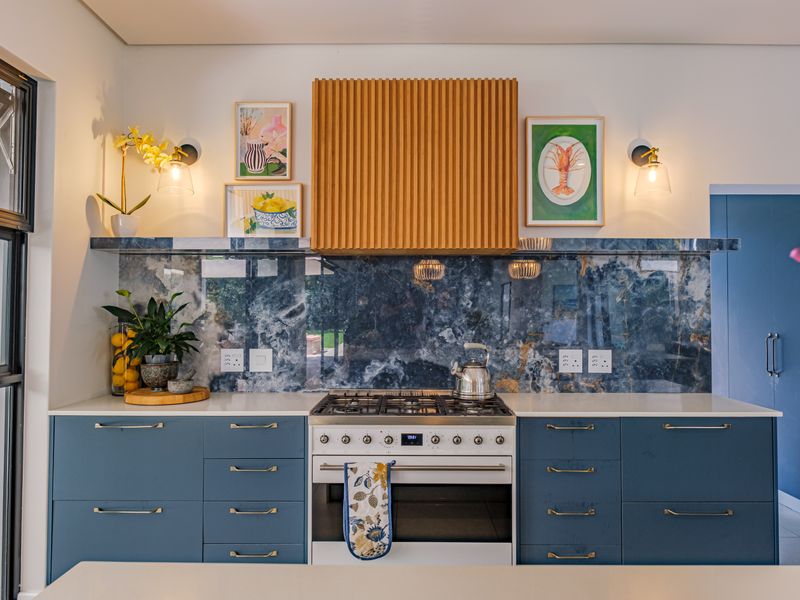Modern kitchen featuring blue cabinetry, a striking stone splashback, a gas hob with a custom timber extractor hood, and sleek stone countertops.