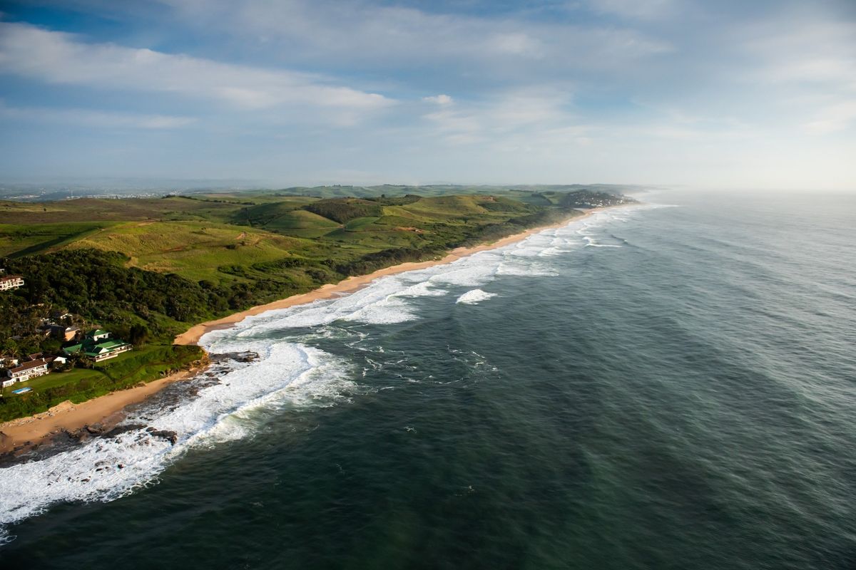 An aerial view of the coastal landscape, showcasing the expansive ocean, sandy beach, and rolling green hills along the shoreline.