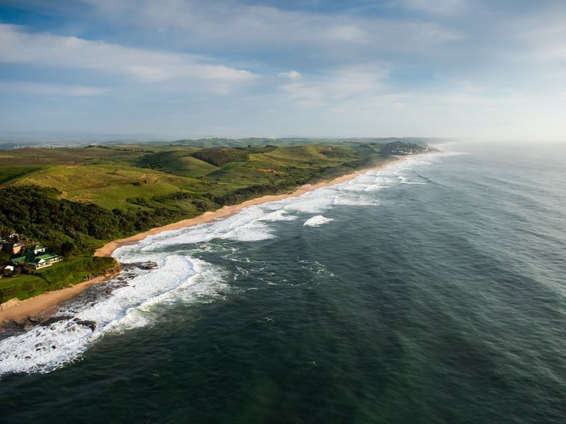An aerial view of the coastal landscape, showcasing the expansive ocean, sandy beach, and rolling green hills along the shoreline.