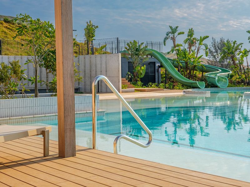 A well-maintained swimming pool area featuring a water slide, timber decking, and modern retaining walls, set within a landscaped residential environment.