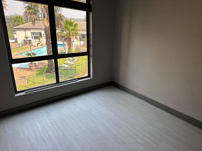 An empty bedroom featuring modern light-toned laminate flooring, neutral walls, and a large window offering a view of the complex pool area.