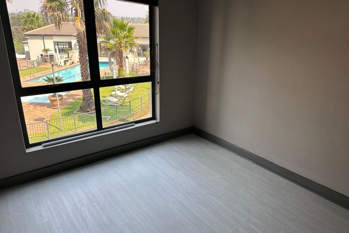 An empty bedroom featuring modern light-toned laminate flooring, neutral walls, and a large window offering a view of the complex pool area.