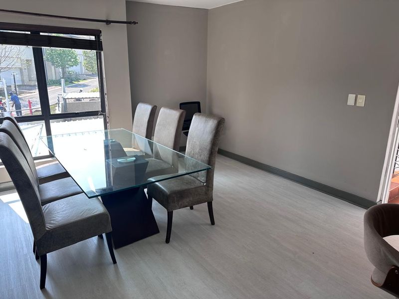 A bright dining area featuring modern light-toned laminate flooring, neutral wall finishes, and a large window providing natural light.
