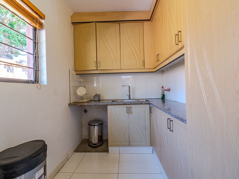 A compact scullery area featuring laminate cabinetry, granite-look countertops, a stainless steel sink, and tiled flooring with security bars on the window.