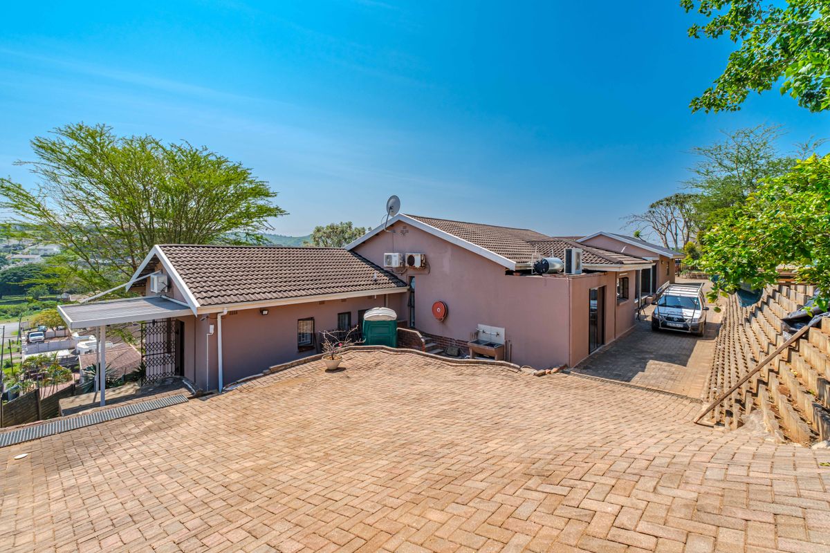 Rear view of the property featuring a paved driveway, tiled roof, and retaining wall structure with external utility fixtures.