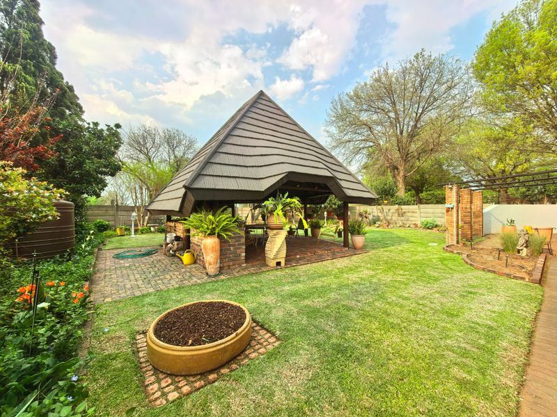 A spacious outdoor entertainment area featuring a covered gazebo with a brick braai, set within a well-maintained garden with brick paving.