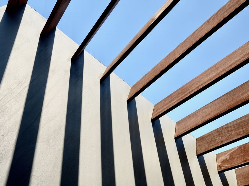 A modern architectural detail featuring a timber pergola structure casting geometric shadows against a clean rendered wall.