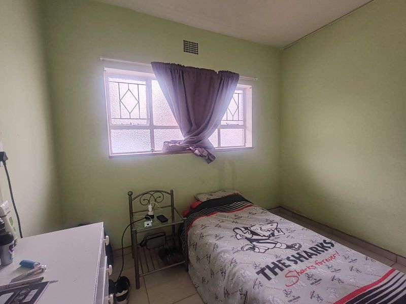 A compact bedroom featuring tiled flooring, a window with security bars, and a ventilation grille, requiring cosmetic updates to the wall finishes.