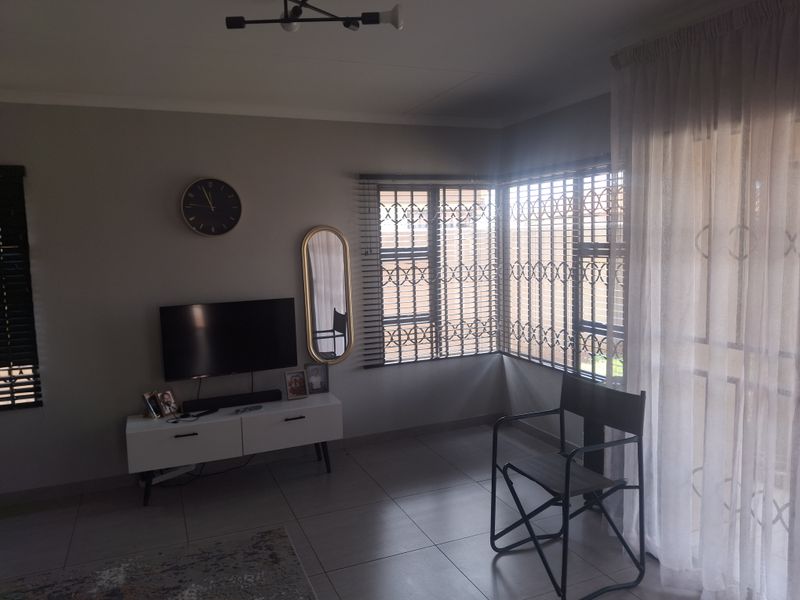 A well-lit living room featuring neutral tiled flooring, large windows with security gates, and modern venetian blinds.