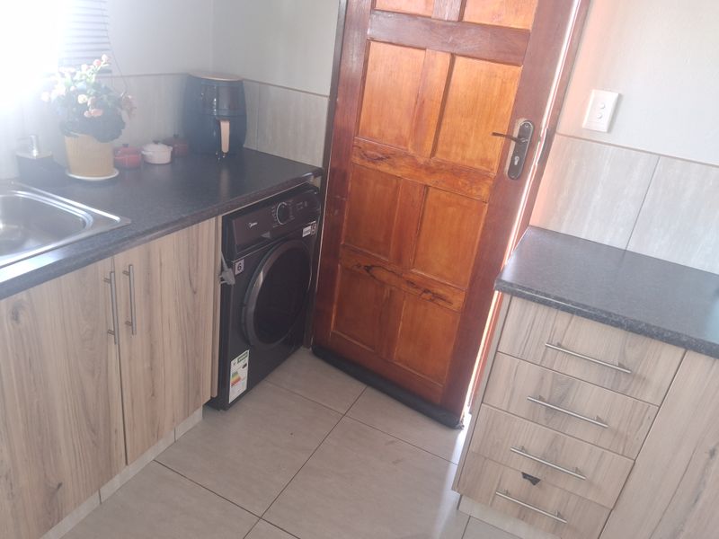 A functional kitchen area featuring light wood-finish cabinetry, dark countertops, a stainless steel sink, and tiled flooring with direct access to an exterior door.