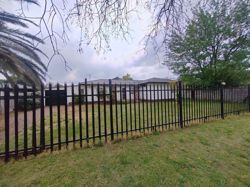 Front view of a single-story residential property featuring a paved driveway, brick facade, and secure metal perimeter fencing.