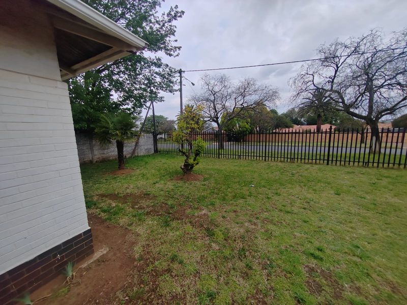 A view of the front yard featuring a painted brick exterior wall, established trees, and metal perimeter fencing.