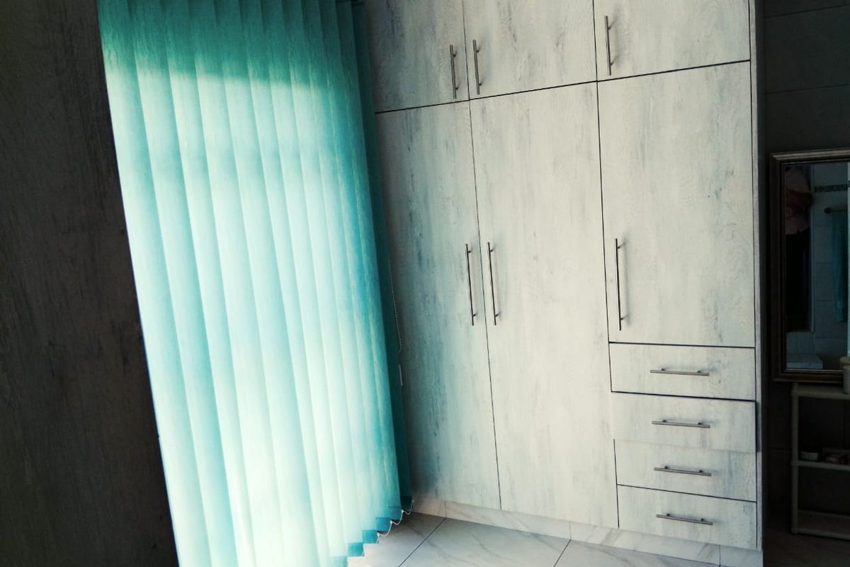 A bedroom featuring light-toned built-in cupboards, tiled flooring, and vertical blinds, offering a clean and functional storage solution.
