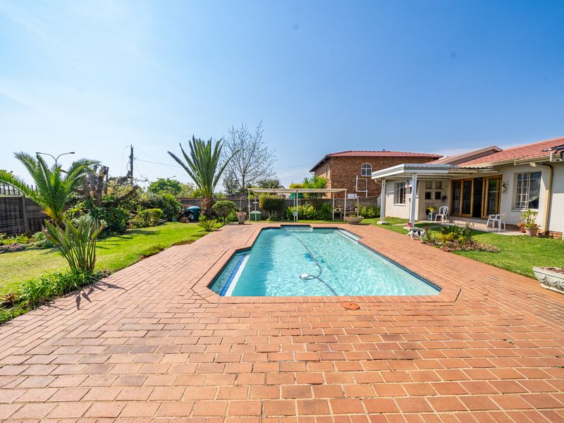 A well-maintained backyard featuring a rectangular swimming pool surrounded by brick paving, a covered patio area, and a landscaped garden.