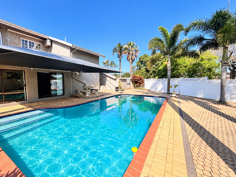 A well-maintained outdoor pool area featuring a paved deck, covered patio space, and secure vinyl perimeter fencing.