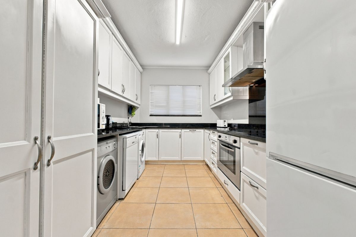 A well-maintained galley-style kitchen featuring white cabinetry, dark granite countertops, built-in oven, extractor fan, and integrated laundry appliances with tiled flooring.
