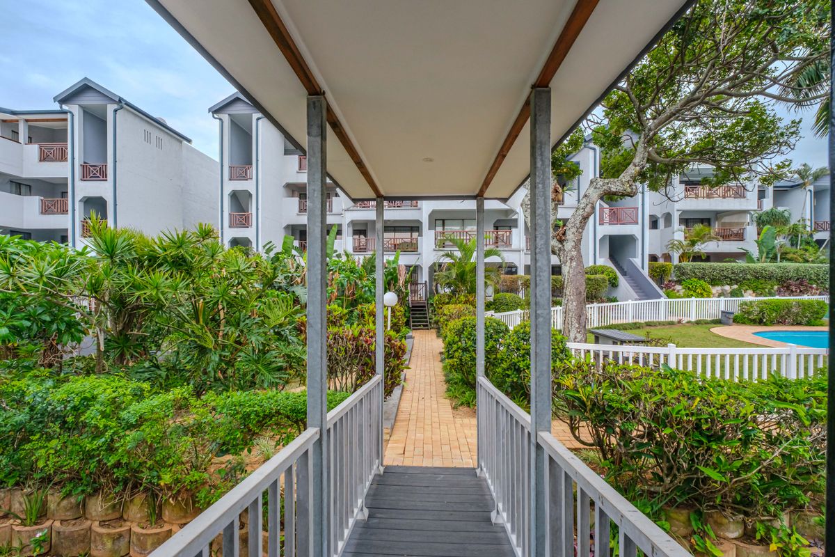 A view of the apartment complex grounds featuring a covered walkway, paved paths, landscaped gardens, and a communal swimming pool area.