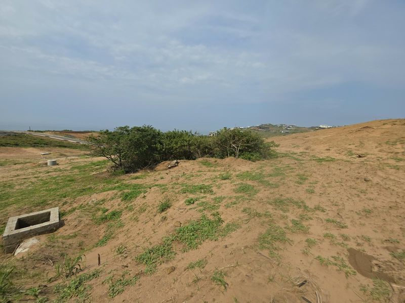 A vacant residential plot featuring natural sandy terrain, native shrubs, and an installed concrete drainage inlet.