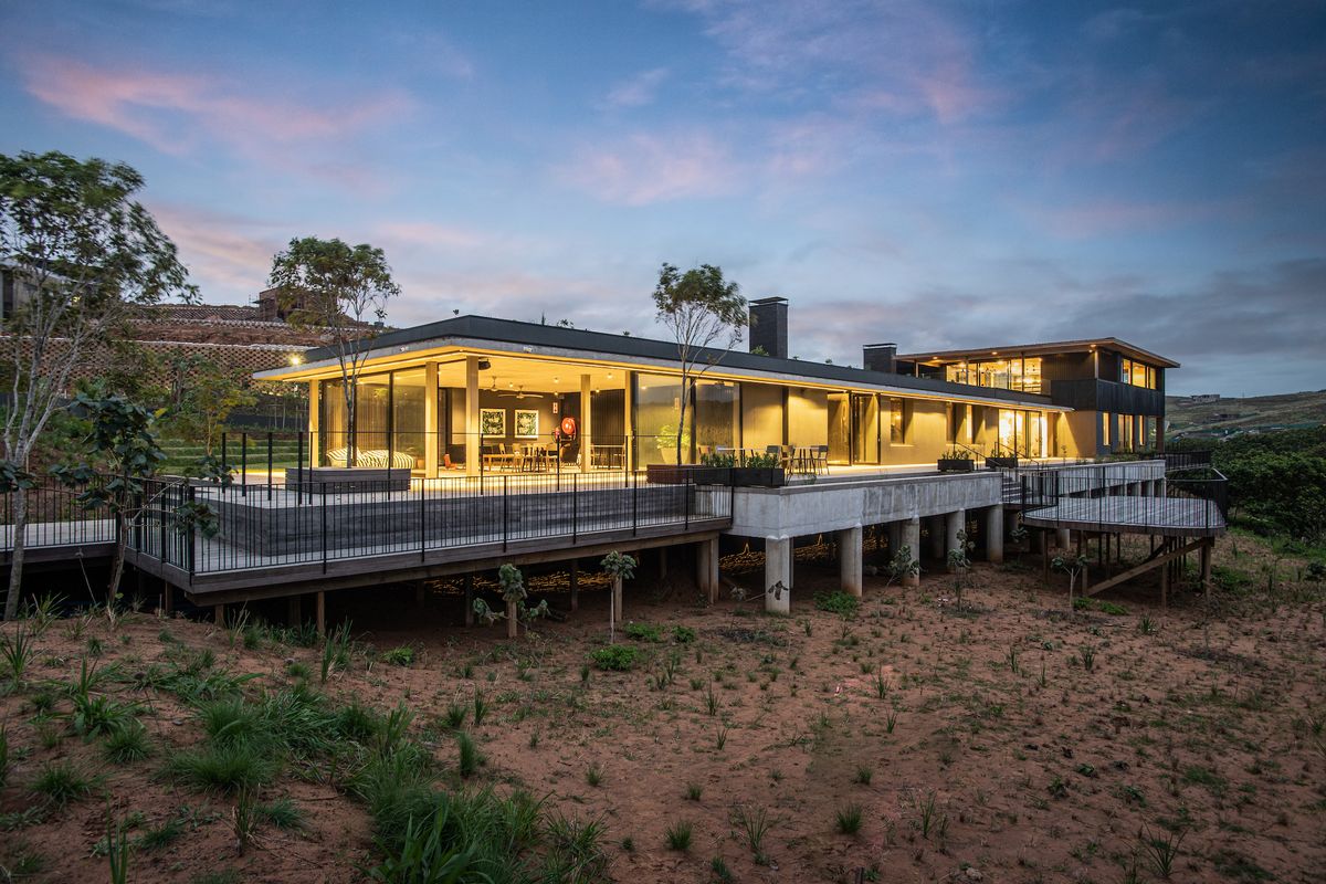Modern architectural residence featuring an expansive raised timber deck, concrete structural supports, and floor-to-ceiling glass walls overlooking a natural landscape.
