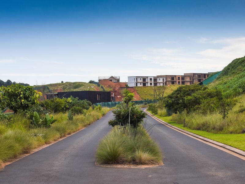 A view of a paved residential street within a development, featuring landscaped verges and surrounding hillside terrain.