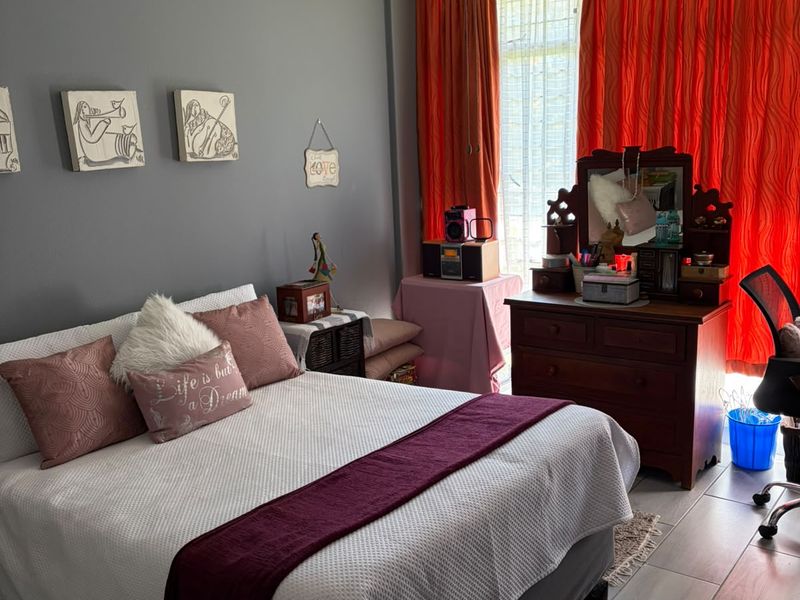 A bedroom featuring tiled flooring and a ceiling fan, with large windows covered by curtains.