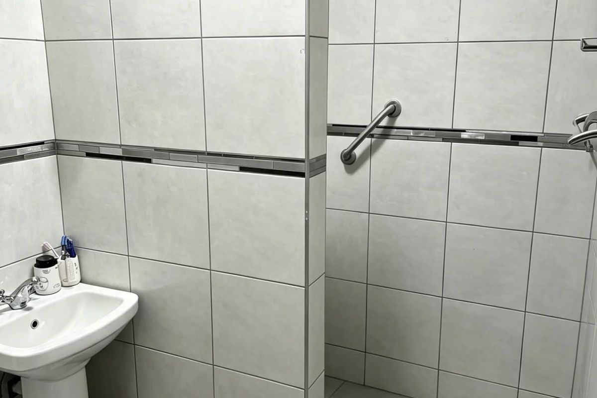 A functional bathroom featuring a pedestal basin, a tiled walk-in shower enclosure with a safety grab rail, and neutral ceramic wall and floor tiling.