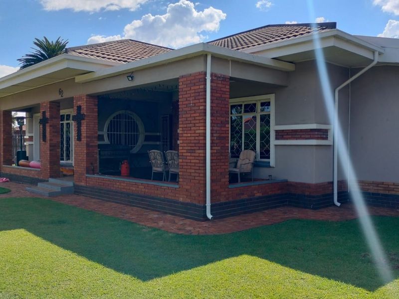 A well-maintained single-story house featuring a covered brick-pillared patio, tiled roof, and neat surrounding lawn.