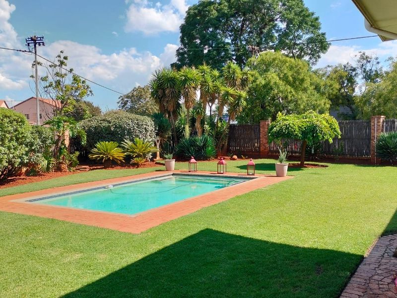 A well-maintained outdoor pool area featuring a clear swimming pool with brick coping, surrounded by a lush green lawn and established garden landscaping.