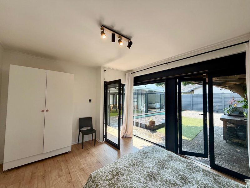 Bright bedroom featuring modern laminate flooring, track lighting, and large black-framed glass doors providing direct access to the outdoor patio and garden area.