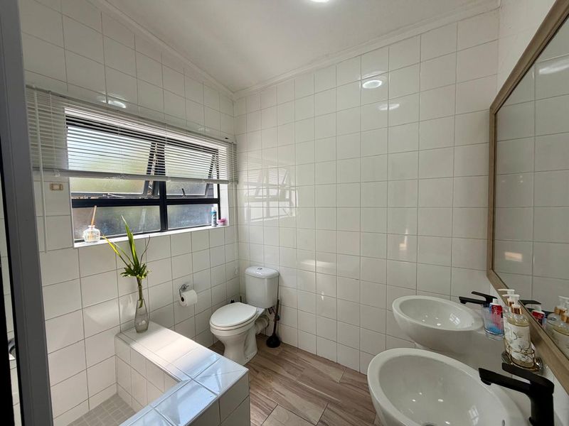 A bright, modern bathroom featuring dual vanity basins, a built-in tiled bathtub, and clean white wall tiling with wood-look flooring.