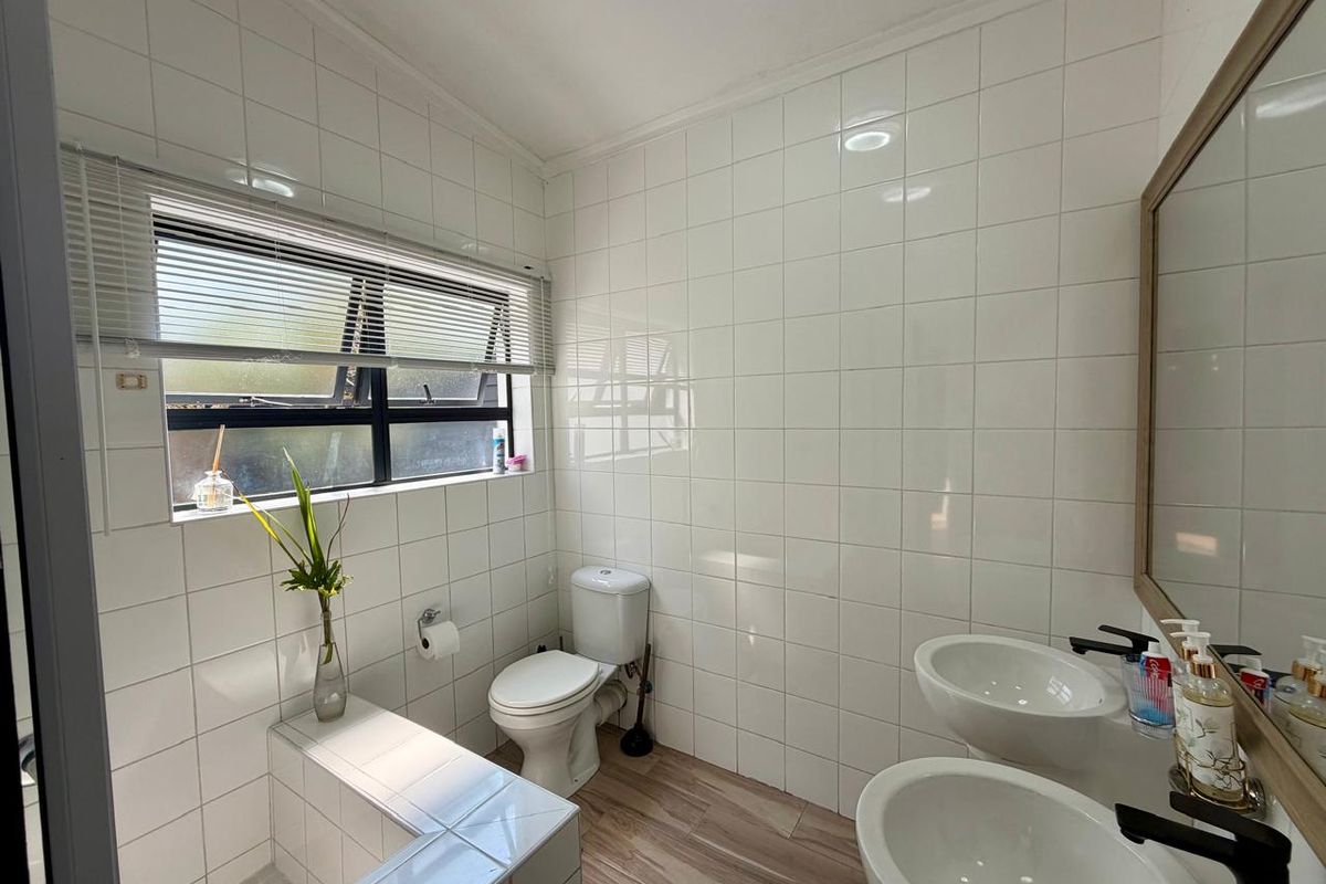A bright, modern bathroom featuring dual vanity basins, a built-in tiled bathtub, and clean white wall tiling with wood-look flooring.