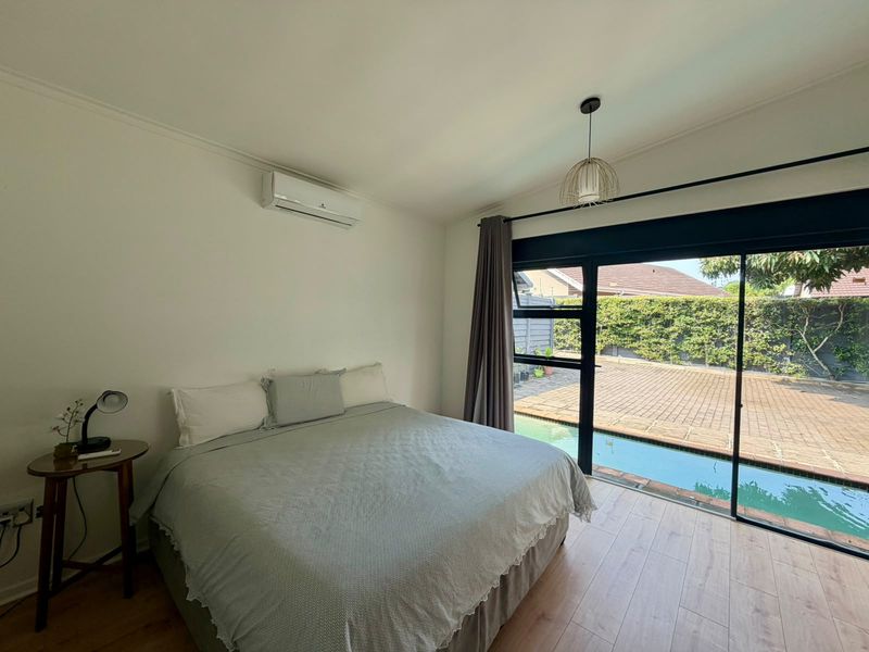 A bright bedroom featuring laminate flooring, a split-system air conditioner, and large sliding glass doors providing direct access to the pool area.