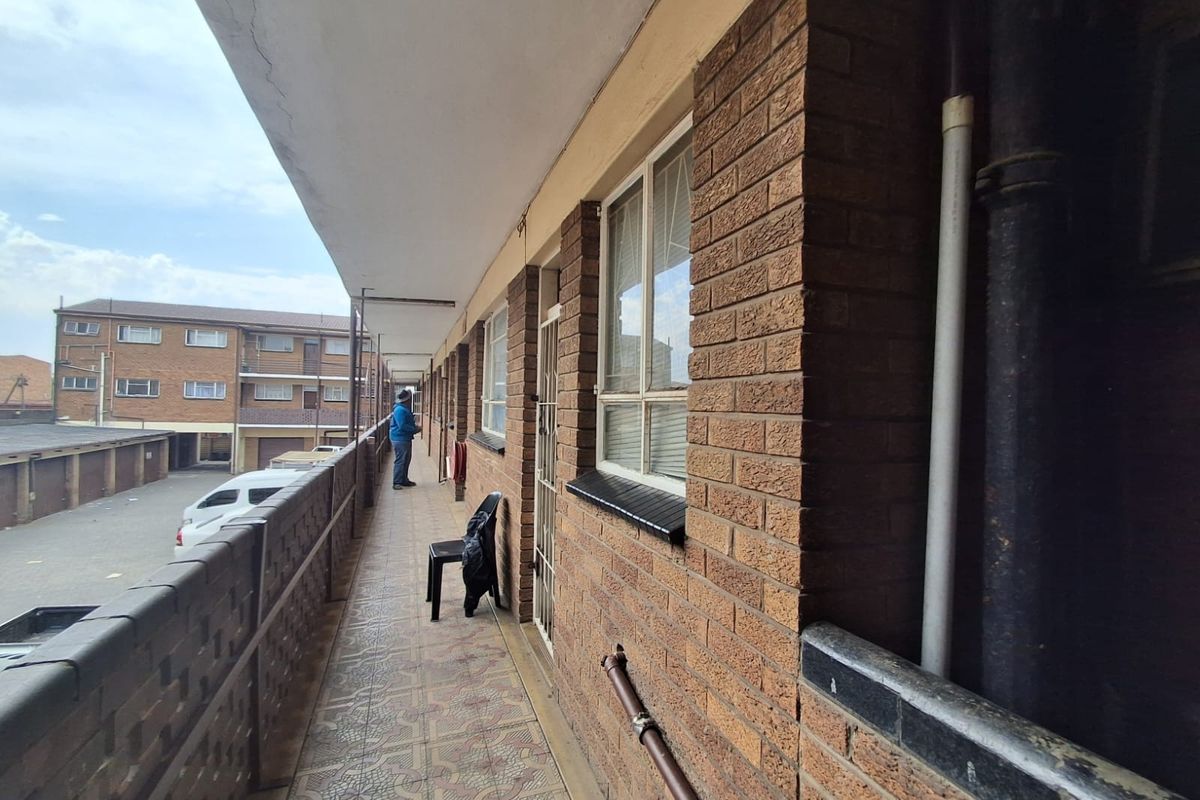 An exterior walkway or balcony featuring brick walls, tiled flooring, and metal balustrades, providing access to the apartment units.