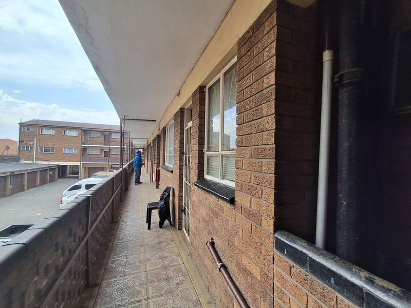 An exterior walkway or balcony featuring brick walls, tiled flooring, and metal balustrades, providing access to the apartment units.