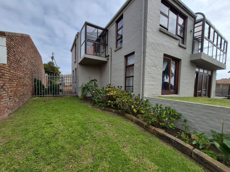 Rear exterior view of a multi-story house featuring a lawn, garden beds, a brick boundary wall, and glass-enclosed balcony structures.