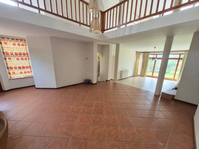 Spacious double-volume living area featuring terracotta floor tiles, structural columns, and a mezzanine level with wooden railing, leading to a garden-facing patio.