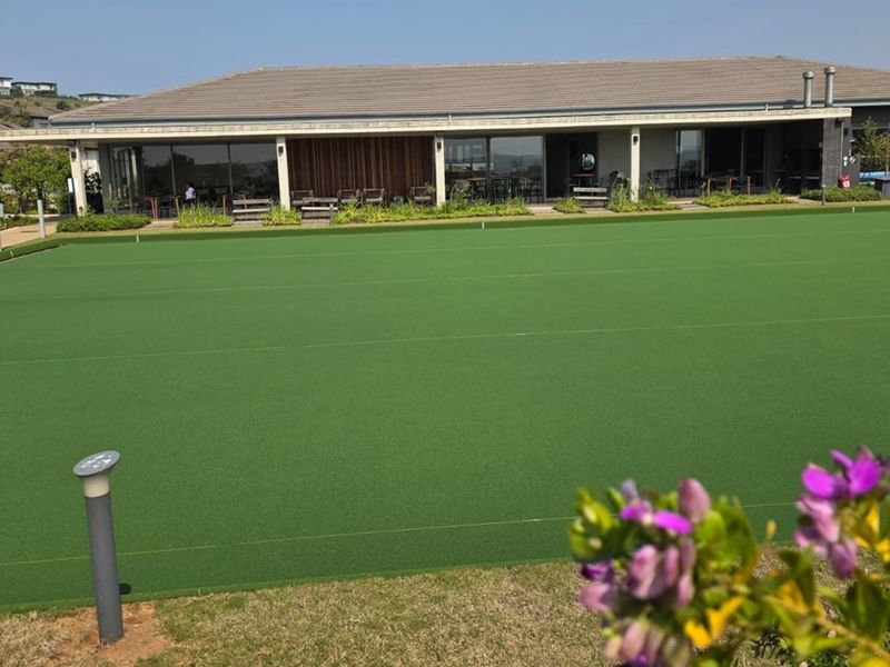 A well-maintained outdoor bowls green situated in front of a modern clubhouse facility with large sliding glass doors.
