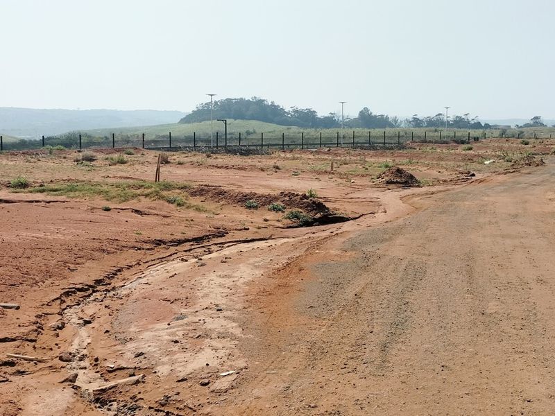 A vacant residential plot featuring unpaved access, perimeter fencing, and distant landscape views, showing signs of soil erosion and uneven terrain.