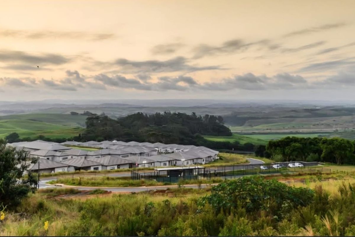 An elevated aerial view of a residential estate featuring paved roads, perimeter security fencing, and surrounding rolling green hills.