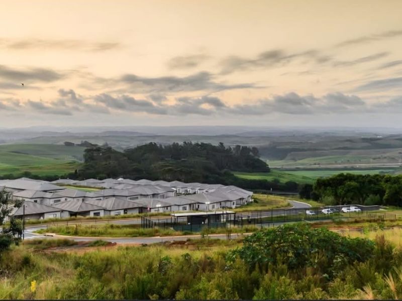 An elevated aerial view of a residential estate featuring paved roads, perimeter security fencing, and surrounding rolling green hills.