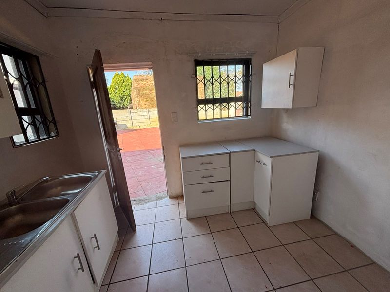 A compact, empty kitchen featuring white built-in cabinetry, a stainless steel sink, tiled floors, and direct access to the rear exterior area.